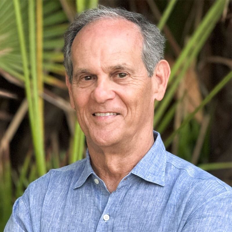 Smiling man standing outdoors with greenery in background.